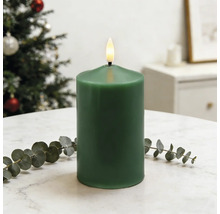Grüne LED-Stumpenkerze aus Echtwachs mit flackernder Flamme auf einem Tisch neben einem Eukalyptuszweig vor einem Weihnachtsbaum.