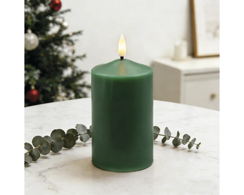 Grüne LED-Stumpenkerze aus Echtwachs mit flackernder Flamme auf einem Tisch neben einem Eukalyptuszweig vor einem Weihnachtsbaum.