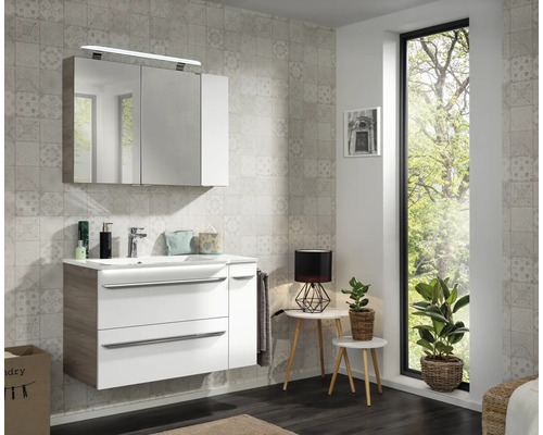 Ensemble de salle de bain blanc avec meuble sous-vasque, vasque en céramique et armoire miroir devant carrelage gris clair, ambiance lumineuse.