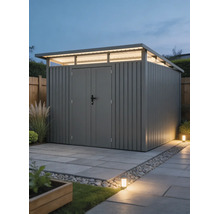 Abri de jardin moderne en métal avec toit en appentis, impostes et double porte sur une surface pavée dans un jardin bien éclairé.