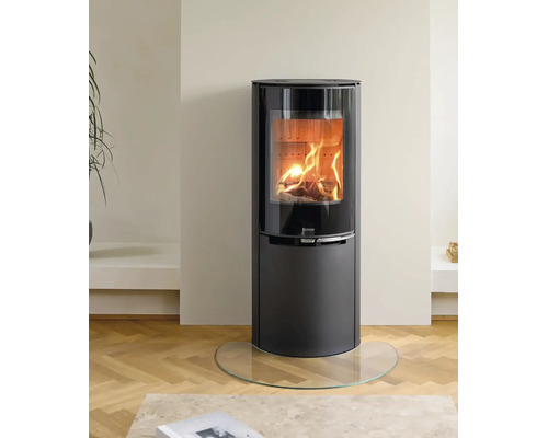 Moderner schwarzer Kaminofen mit brennendem Holzfeuer hinter einer großen Glasscheibe auf einer Glasbodenplatte in einem hellen Wohnraum.
