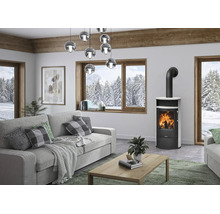 Salon moderne avec poêle à bois autoportant en acier noir avec habillage blanc, canapé gris, table basse en bois et grandes fenêtres.