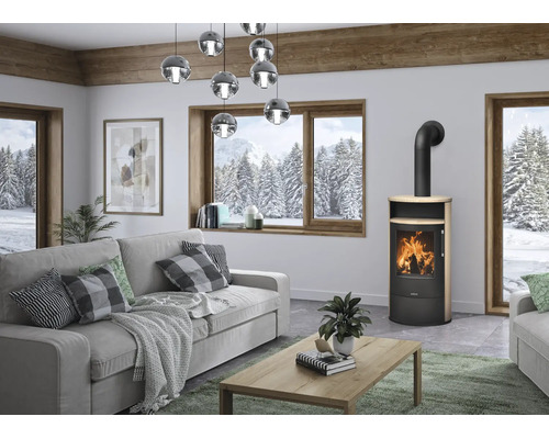 Poêle à bois moderne en métal noir et bois dans un salon lumineux avec un canapé gris et de grandes fenêtres donnant sur les montagnes.
