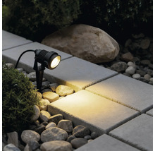 Spot de jardin LED noir avec piquet éclairant une surface pavée entre des pierres et des plantes avec une lumière blanc chaud.
