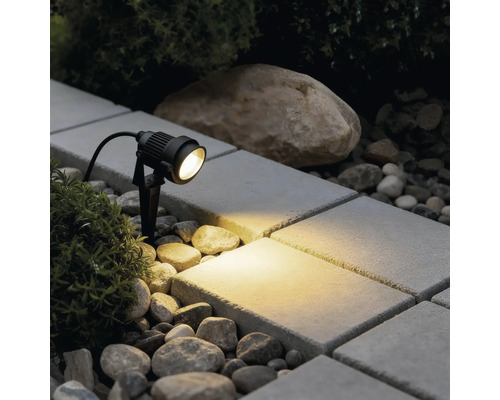 Spot de jardin LED noir avec piquet éclairant une surface pavée entre des pierres et des plantes avec une lumière blanc chaud.
