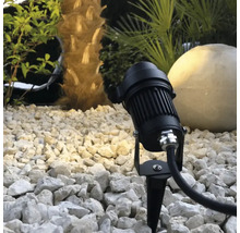 Spot de jardin noir avec piquet de terre dans un lit de gravier devant un palmier et une boule lumineuse.