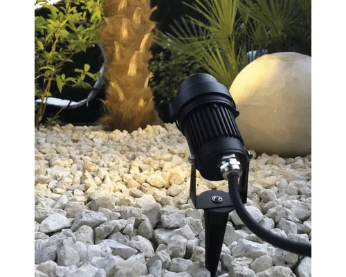 Spot de jardin noir avec piquet de terre dans un lit de gravier devant un palmier et une boule lumineuse.
