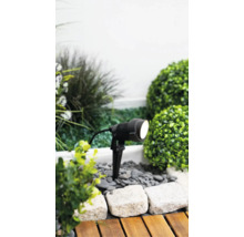 Spot de jardin à LED noir avec piquet de terre dans un parterre de gravier et de plantes à côté d'une terrasse en bois.