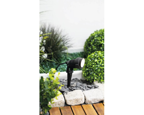 Spot de jardin à LED noir avec piquet de terre dans un parterre de gravier et de plantes à côté d'une terrasse en bois.