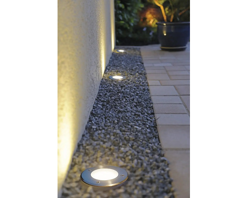 Spots encastrés de sol LED extérieurs dans un lit de gravier le long d'un mur, éclairant vers le haut avec une lumière blanche chaude.