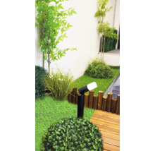 Scène de jardin moderne avec un spot extérieur LED noir, du gazon synthétique, des plantes vertes, une bordure en bois et une terrasse en bois.