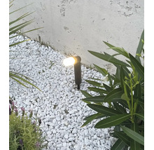 Spot de jardin éclairé avec piquet dans un lit de gravier entre des plantes devant un mur structuré.