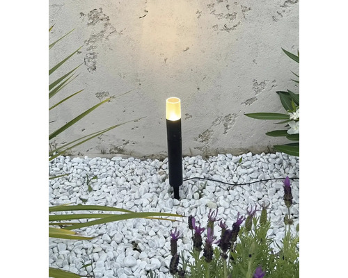 Lampe de jardin noire avec piquet dans un lit de gravier blanc devant un mur texturé.