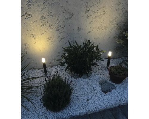 Deux spots de jardin à LED avec piquet dans un parterre de gravier et diverses plantes devant un mur crépi.
