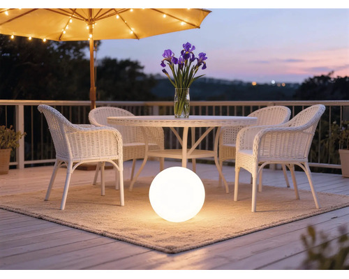 Lampe sphérique éclairée sur un tapis sous un mobilier de jardin en rotin blanc sur une terrasse organisée au crépuscule.