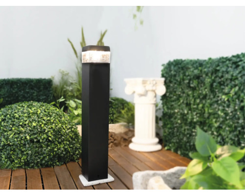 Borne d éclairage extérieur noire moderne sur une terrasse en bois dans un jardin, entourée de plantes vertes.