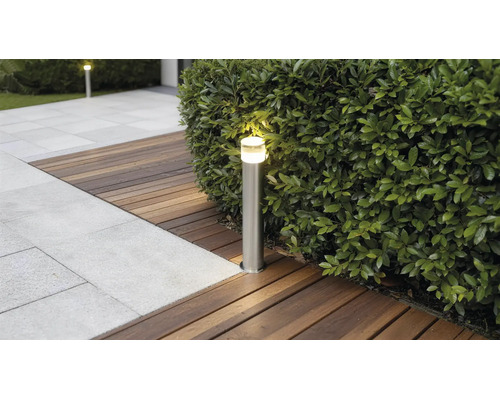 Borne d'éclairage extérieur éclairée en acier inoxydable sur une terrasse en bois à côté d'une haie et d'un chemin en pierre.