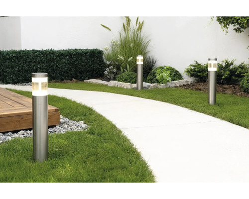 Trois bornes lumineuses modernes en acier inoxydable éclairant une allée de jardin courbée à côté de la pelouse et des massifs.
