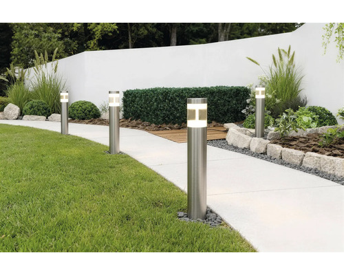 Bornes lumineuses modernes en acier inoxydable éclairant une allée de jardin claire à côté d'une pelouse et de parterres bordés de pierres.