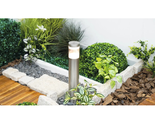 Borne lumineuse cylindrique en acier inoxydable dans un parterre de jardin moderne avec pierres et plantes.