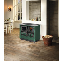Cuisinière à bois Justus en vert foncé avec feu allumé dans une cuisine lumineuse avec sol en bois et panier de bois de chauffage.