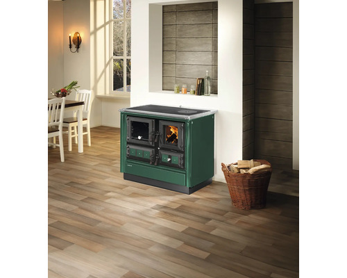 Cuisinière à bois verte dans une cuisine lumineuse avec parquet, table à manger et panier de bois de chauffage.