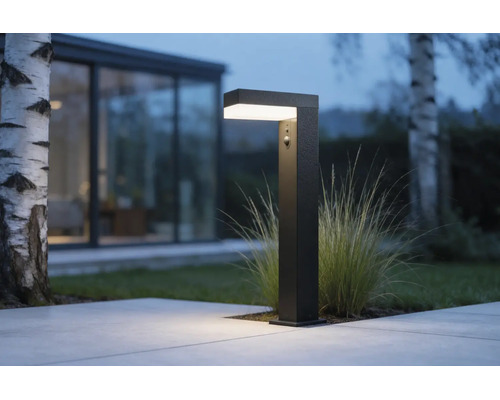 Borne lumineuse LED noire avec détecteur de mouvement éclairant un chemin de terrasse devant une maison moderne au crépuscule.