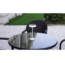 Lampe de table LED moderne sur une table ronde en verre dans un jardin avec des chaises en rotin foncé et du gravier blanc.