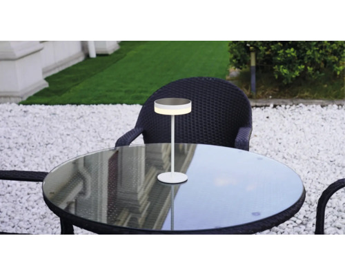 Lampe de table LED moderne sur une table ronde en verre dans un jardin avec des chaises en rotin foncé et du gravier blanc.