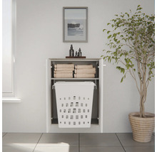 Armoire de salle de bain blanche avec panier à linge intégré et étagère pour serviettes, fixée sur un mur clair à côté d'une plante.