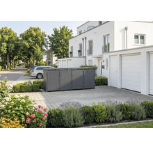 Moderne Mülltonnenbox aus Metall für vier Tonnen in einer gepflasterten Einfahrt vor einem weissen Wohnhaus mit Bepflanzung.