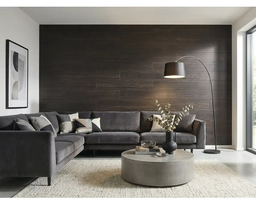 Salon moderne avec revêtement mural sombre aspect bois, canapé d'angle gris en velours, table basse ronde en béton et lampadaire arqué.