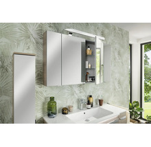 Armoire de toilette à trois portes avec éclairage dans la salle de bain, montée au-dessus d'un lavabo blanc devant un papier peint à motifs.