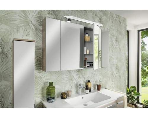 Armoire de toilette à trois portes avec éclairage dans la salle de bain, montée au-dessus d'un lavabo blanc devant un papier peint à motifs.