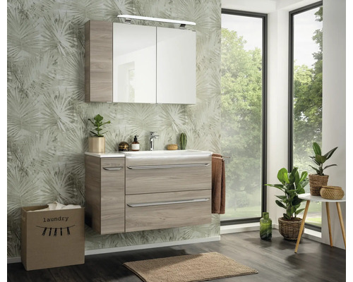 Modernes Badezimmer-Set mit Waschtischunterschrank in Holzoptik, weißem Waschbecken und Spiegelschrank vor Palmentapete.