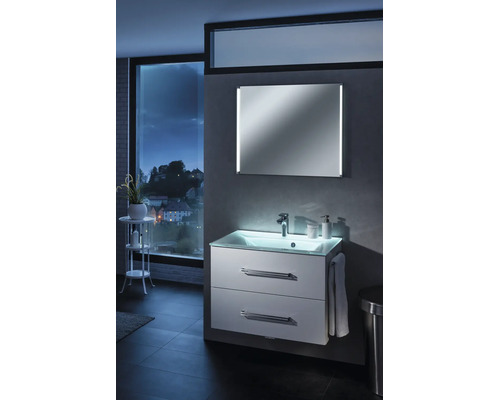 Meuble sous-vasque moderne blanc éclairé avec plan de toilette en verre et miroir dans une salle de bains avec vue sur la ville la nuit.