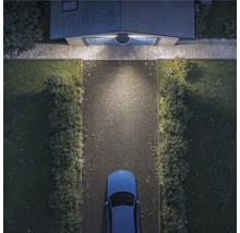 Vogelperspektive einer hell beleuchteten Einfahrt bei Nacht mit Außenleuchte über dem Garagentor, Schotterweg, Hecken und Auto.