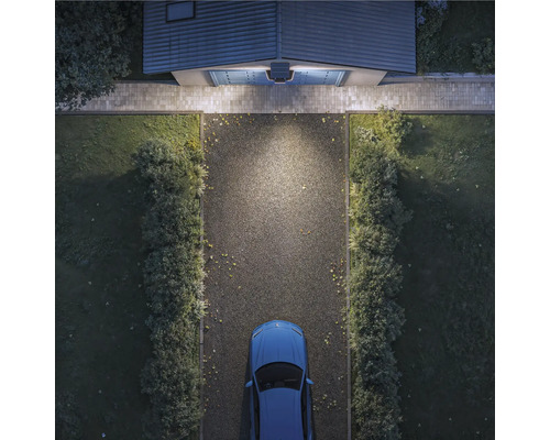 Vogelperspektive einer hell beleuchteten Einfahrt bei Nacht mit Außenleuchte über dem Garagentor, Schotterweg, Hecken und Auto.