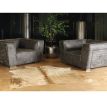 Deux fauteuils en cuir gris avec des coutures décoratives sur un parquet, un tapis en peau de vache et une plante verte dans un pot en métal.