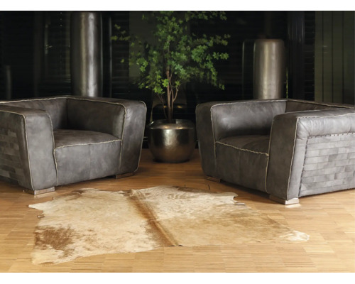Deux fauteuils en cuir gris avec des coutures décoratives sur un parquet, un tapis en peau de vache et une plante verte dans un pot en métal.