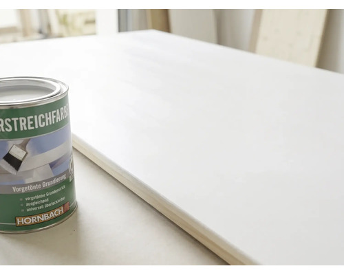 Eine Dose Hornbach Vorstreichfarbe neben einer Holzplatte, die mit einer weißen Grundierung gestrichen wurde.