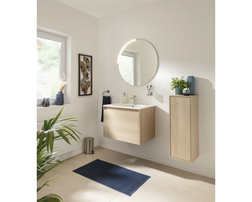 Salle de bains moderne et lumineuse avec meuble sous-vasque suspendu aspect bois, miroir rond, armoire latérale et tapis de bain bleu foncé.