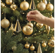 Eine Hand hängt eine glänzende, runde Weihnachtskugel an einen geschmückten Weihnachtsbaum mit verschiedenen Ornamenten und einer Lichterkette.
