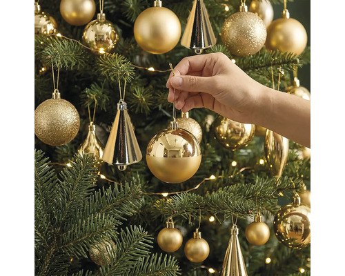 Eine Hand hängt eine glänzende, runde Weihnachtskugel an einen geschmückten Weihnachtsbaum mit verschiedenen Ornamenten und einer Lichterkette.