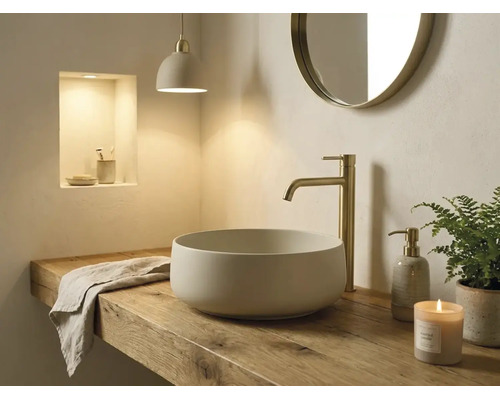 Helles Badezimmer mit rundem Aufsatzwaschbecken auf einer Holzplatte, einer hohen goldfarbenen Armatur und einem runden Wandspiegel.