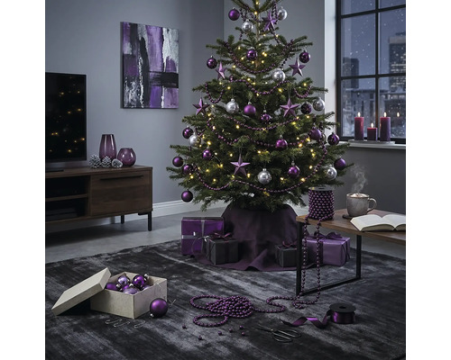 Festlich geschmückter Weihnachtsbaum mit violetten und silbernen Kugeln sowie Sternen in einem modern eingerichteten Wohnzimmer mit Geschenken.