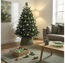 Dekorierter künstlicher Weihnachtsbaum mit grünen Kugeln in verschiedenen Grössen in einem geflochtenen Ständer im hellen Wohnzimmer.