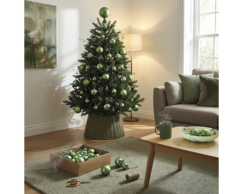 Dekorierter künstlicher Weihnachtsbaum mit grünen Kugeln in verschiedenen Grössen in einem geflochtenen Ständer im hellen Wohnzimmer.