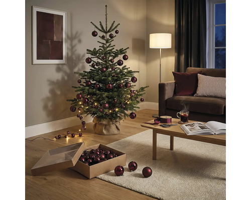 Festliches Wohnzimmer mit einer Nordmanntanne, geschmückt mit dunkelroten Weihnachtskugeln und LED-Lichterkette, neben Sofa und Couchtisch.