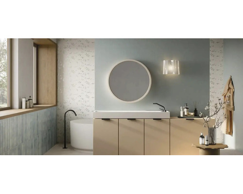 Modernes Badezimmer mit beigem Waschtischunterschrank, weißem Waschbecken, rundem Spiegel und freistehender Badewanne vor floralen Fliesen.